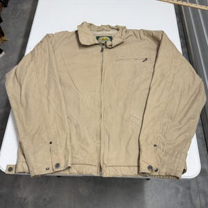 Puede incluir: Una chaqueta de lona color canela con cremallera frontal completa, cuello vuelto y una etiqueta de la marca Cabela's en el interior del cuello. La chaqueta incluye un bolsillo en el pecho con cremallera, dos bolsillos inferiores y puños.