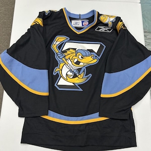 May include: A black Reebok hockey jersey featuring a large graphic of a yellow fish holding a hockey stick over a white letter F. The jersey has light blue and yellow stripes on the sleeves and hem, with a Reebok logo and ECHL patch on the collar.