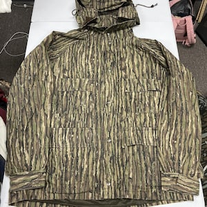 May include: A long-sleeve hooded jacket featuring a tree bark camouflage pattern in shades of brown, green, and black. The jacket includes a front button closure, two large chest pockets with flaps, and adjustable cuffs with hook-and-loop fasteners.