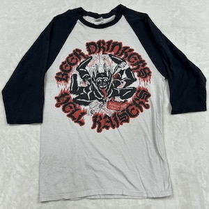 May include: A light gray baseball t-shirt with black three-quarter length sleeves. The front features a graphic of a multi-armed creature with a boombox and beer, surrounded by the red text BEER DRINKERS HELL RAISERS in a circular design.