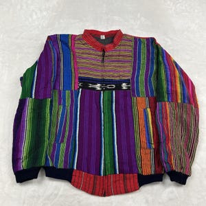 May include: A zip-up jacket featuring a vibrant, multi-colored striped pattern in shades of purple, green, blue, red, and orange. The garment includes a horizontal black band with white geometric motifs across the chest and dark cuffs.