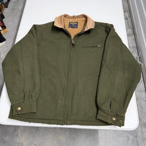Op de afbeelding: Een olijfgroene jas van wolmix met een beige corduroy kraag, een ritssluiting over de gehele lengte, een borstzak met rits en lange mouwen met knoopmanchetten. Het label aan de binnenzijde toont de merknaam Woolrich in blauw en goud.