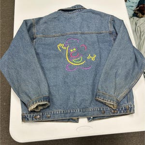 May include: A blue denim jacket featuring a large, colourful embroidered graphic of Mr. Potato Head on the back. The embroidery uses neon yellow, pink, and green thread to outline the character, which includes a hat, moustache, and wide smile.