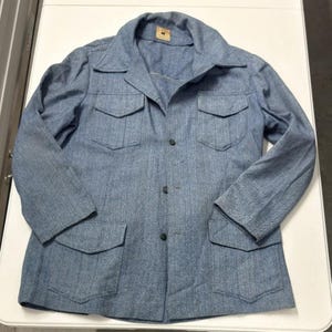 Può includere: Giacca di jeans blu chiaro con quattro tasche frontali e colletto. La giacca ha una chiusura con bottoni e maniche lunghe. Una piccola etichetta è visibile vicino al colletto. La giacca è realizzata in tessuto strutturato.