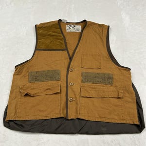 May include: A tan canvas vest featuring a dark brown quilted shoulder patch, a dark brown lower back panel, and multiple utility pockets. The front includes three buttons, a chest pocket, and a Duck Bay brand label with a duck illustration.