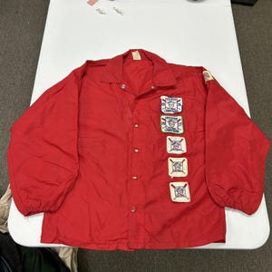May include: A bright red long-sleeve windbreaker jacket featuring a pointed collar, a snap-button front closure, and elastic cuffs. The front is decorated with five embroidered patches related to softball umpire certification from 1976 and 1977.