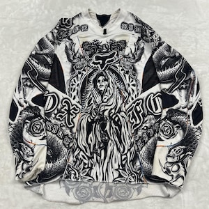 May include: A long-sleeve jersey featuring a black and white graphic print of a hooded figure with hands in prayer, surrounded by roses, snakes, and the Fox Racing logo. The design includes the text "LIVE RIDE" and "FOX" in a gothic font.