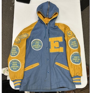 May include: A light blue and yellow varsity jacket with a hood, featuring a large yellow letter E on the chest, yellow leather sleeves, and multiple circular patches detailing sports achievements like AAAA State Runner-Up and Region Champions.