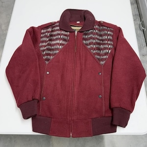 May include: A burgundy wool jacket with a full zipper closure. The jacket features a patterned design on the upper chest and shoulders, with a ribbed collar and cuffs. The jacket has two small buttons on each side.