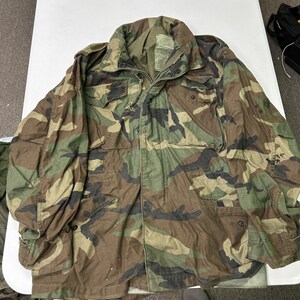 May include: A camouflage field jacket featuring a woodland pattern in shades of green, brown, and black. The jacket includes a fold-down collar, a front zipper closure, two chest pockets with flaps, and two lower pockets with snap button closures.