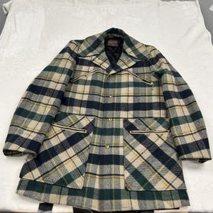 May include: A Pendleton plaid jacket featuring a tan, dark green, and navy blue checkered pattern. The jacket includes a collared neckline, a front button closure, two zippered chest pockets, and two large zippered pockets at the waist.