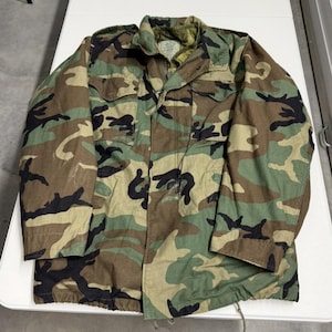 Puede incluir: Chaqueta de camuflaje en tonos verdes, marrones y negros. La chaqueta tiene cierre de cremallera, varios bolsillos y un cordón en la parte inferior. La chaqueta está hecha de tela resistente. Estilo militar clásico.
