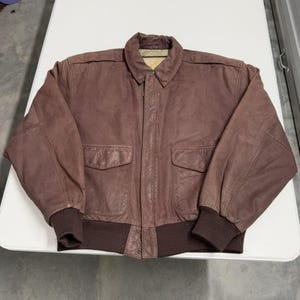 Könnte beinhalten: Braune Leder-Bomberjacke mit Kragen, zwei Vordertaschen und gerippten Bündchen und Saum. Die Jacke hat ein klassisches Design und eine Vintage-Ästhetik. Das Leder wirkt abgenutzt, was zu seinem Charakter beiträgt.