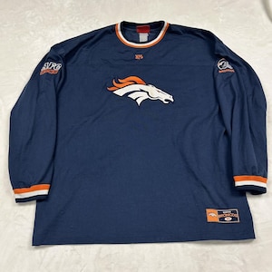 May include: A navy blue long-sleeve shirt featuring the Denver Broncos logo on the chest. The shirt has orange and white striped cuffs and collar, an NFL logo at the neck, an AFC West patch on the right sleeve, and a Broncos patch on the left.