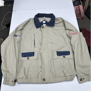 May include: A beige jacket with a dark blue collar, featuring a vertical zippered chest pocket, two large front flap pockets with buttons, and long sleeves. The right sleeve displays a Wyeth-Ayerst logo, and the left sleeve features a red Coca-Cola logo.