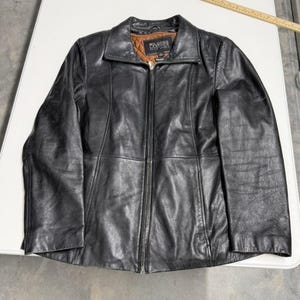 Puede incluir: Chaqueta de cuero negra con cremallera, cuello y etiqueta de la marca "WILSONS LEATHER". La chaqueta tiene un forro marrón acolchado y está colocada sobre una superficie blanca. Una regla de madera está en el fondo.