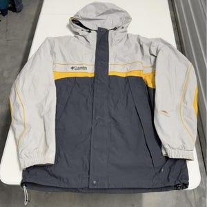 Puede incluir: Chaqueta con capucha Columbia gris, amarilla y azul marino. La chaqueta tiene capucha y mangas grises, una franja amarilla en el pecho y una sección inferior azul marino. El logotipo de Columbia es visible.