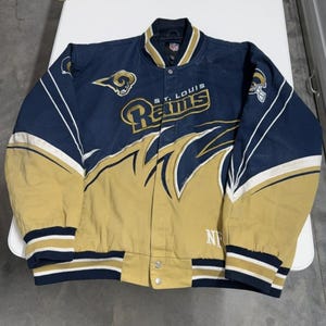 Puede incluir: Una chaqueta de satén azul marino y dorado de los Rams de St. Louis. La chaqueta presenta el logotipo del equipo y las palabras "St. Louis Rams" en letras doradas. La chaqueta tiene cierre de botones y detalles en blanco.