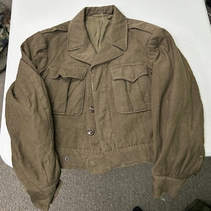 May include: A khaki-coloured wool military-style jacket featuring a collared neckline, two large button-flap chest pockets, and a front button closure. The garment includes long sleeves with cuffs, shoulder epaulets, and a waist adjustment strap.