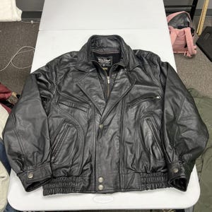 May include: A black leather jacket featuring a fold-down collar, a full-length front zipper, and a snap-button closure. The jacket includes two zippered chest pockets, two lower zippered pockets, and a ribbed elastic hem. A Wilsons label is visible.