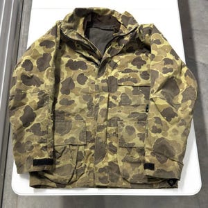 May include: A camouflage jacket with a duck pattern in shades of brown, tan, and olive green. The jacket has a zip-up front, multiple pockets, and a high collar. The sleeves have adjustable cuffs.
