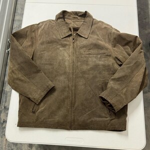 May include: A brown suede jacket featuring a full-length front zipper, a standard collar, long sleeves, and two lower zippered pockets. The garment includes a single horizontal zippered chest pocket on the left side and a textured finish.