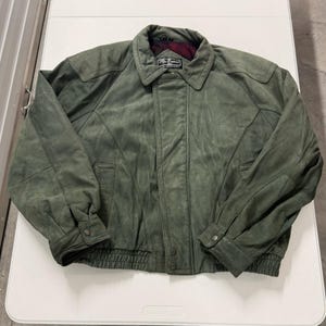 May include: A green suede jacket with a button-up front, two front pockets, and a collared neckline. The jacket has a ribbed hem and cuffs. The interior collar lining is a red and black plaid pattern. The brand name "Peter Shepherd" is visible on the collar.