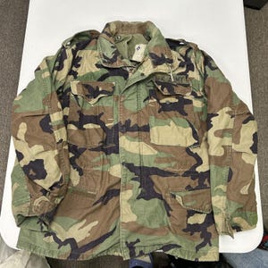 May include: A long-sleeve field jacket featuring a woodland camouflage pattern in shades of green, brown, and black. The jacket includes a fold-down collar, shoulder epaulets, four front pockets with button flaps, and a U.S. ARMY patch.