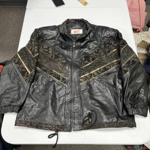 May include: A black leather jacket featuring a front zipper closure, a high collar, and long sleeves with elastic cuffs. The jacket includes dark patterned fabric panels across the chest and hem, accented with gold-toned piping and two drawstring cords.