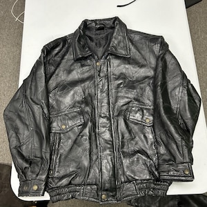 Puede incluir: Una chaqueta de cuero negro confeccionada con múltiples paneles cosidos. Presenta un cuello clásico, cierre frontal de cremallera con tapeta abotonada, dos bolsillos delanteros con solapa y cierre de botón, puños abotonados y un dobladillo elástico.