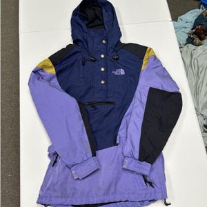 May include: A color-blocked pullover jacket featuring a navy blue chest and hood, light purple sleeves and lower body, and black shoulder and arm accents. The jacket includes a front zipper pocket, a snap-button placket, and The North Face logo.
