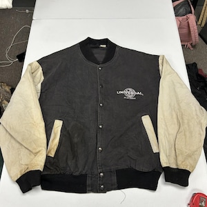 May include: A black jacket with off-white sleeves, a black ribbed collar, and black ribbed cuffs. The front features a button-down closure, two side pockets with off-white trim, and the white embroidered text Universal An MCA Company.