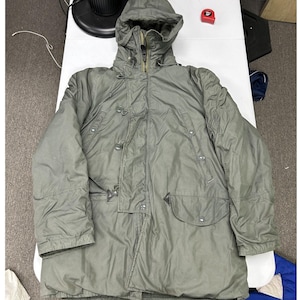 May include: An olive green hooded parka featuring a full-length front zipper, a button-down storm flap, and two large front pockets with button closures. The jacket includes a drawstring hood and long sleeves, shown with a red measuring tape.