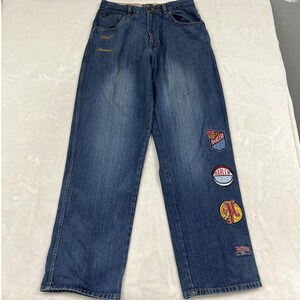 May include: Medium-wash blue denim jeans featuring a button fly and five-pocket construction. The left leg displays three circular embroidered patches with Harlem-themed graphics and text, plus a small rectangular patch near the hem.