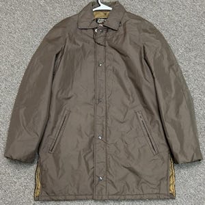 May include: A brown long-sleeve jacket with a collar, a front button closure, and two zippered side pockets. The jacket features a visible interior quilted lining in a tan colour and a label at the neck that reads Oakbrook Sportswear.