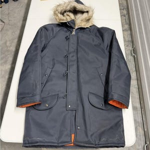 Op de afbeelding: Een marineblauwe winterparka met een met imitatiebont gevoerde capuchon. De jas heeft een ritssluiting en knevelsluitingen, twee voorzakken en een oranje voering zichtbaar bij de manchetten en zoom. Dit bovenkledingstuk is ontworpen voor koud weer.