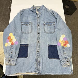 May include: A light blue denim shirt featuring a button-down front, two chest pockets, and two dark blue patch pockets at the waist. Each sleeve is decorated with a cluster of multicolored, hexagonal fabric patches in yellow, orange, and purple.
