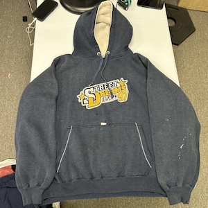 Könnte beinhalten: Ein marineblauer Kapuzenpullover mit cremefarbenem Innenfutter und Kapuzenkordel. Auf der Vorderseite befindet sich ein Aufdruck mit dem Text "STREET DREAMS IB N.Y.C 97" in gelber und weißer Schrift, akzentuiert durch gelbe Sterne und weiße Nähte.