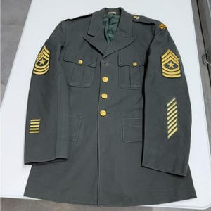 Op de afbeelding: Donkergroene militaire uniformjas met gouden knopen en emblemen. De jas heeft meerdere zakken, schouderstukken en sergeantstrepen op de mouwen. De stof is duurzaam en maakt deel uit van een militair uniform.