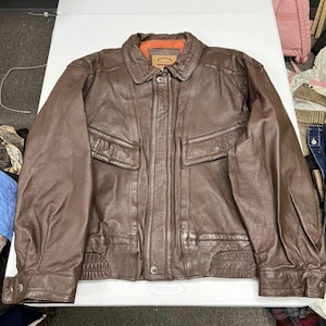 May include: A brown leather jacket featuring a classic collar, long sleeves with buttoned cuffs, and two front chest pockets with snap closures. The jacket includes a central button placket and a bright orange quilted interior lining visible at the neck.