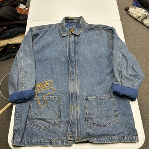 May include: A blue denim jacket featuring a fold-down collar, long sleeves with rolled cuffs, and a front button closure. The jacket includes two large patch pockets on the front and an embroidered yellow Fila logo on the right chest area.
