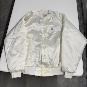 May include: Cream-colored satin bomber jacket with a button-up front, ribbed cuffs, and a collar. The jacket features the Motorola logo embroidered in blue on the chest. A vintage-style jacket.