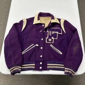 Puede incluir: Una chaqueta universitaria morada con cuello, detalles en los hombros y ribetes de bolsillo en color crema. El frente muestra una letra F grande de estilo bloque en morado con contorno crema. Tiene cierre de botones a presión y puños acanalados.