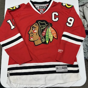 May include: A red Reebok Chicago Blackhawks hockey jersey featuring a large team logo on the chest, the number 19 on the sleeves, and the number 9 with a captain's C on the front. The jersey includes black and white stripes at the hem and sleeves.