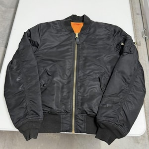May include: Black bomber jacket with a gold zipper and orange lining. The jacket has two front pockets, a ribbed collar, cuffs, and waistband. The jacket is made of a smooth, shiny material.