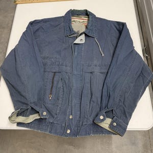 May include: A blue cotton jacket featuring a collared neckline, a front zipper closure with a snap-button placket, long sleeves with snap-button cuffs, and a patterned interior lining visible at the collar. The jacket includes a zippered chest pocket.