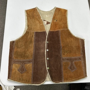 Pode incluir: Um colete sem mangas feito de retalhos de camurça em vários tons de castanho, com forro de pele sintética creme, decote em V, quatro botões de pressão de metal prateado e dois bolsos frontais decorativos.
