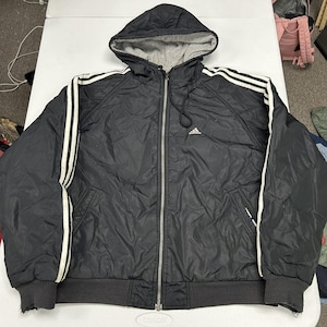 May include: A black zip-up hooded jacket featuring a grey interior lining, a full-length front zipper, and three white stripes running down the length of each sleeve. The Adidas logo is embroidered in white on the upper left chest area.