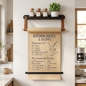 May include: A wall-mounted paper roll dispenser with a black metal shelf and tan leather straps. The kraft paper features printed text: KITCHEN NOTES & RECIPES, a shopping list, and a sourdough recipe with illustrations of kitchen tools.