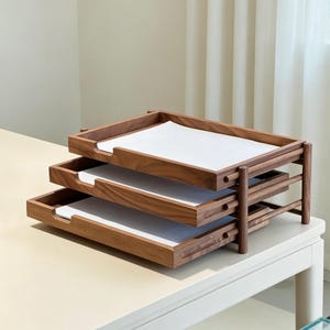 May include: A three-tiered wooden desk organiser with a warm brown finish. Each tray holds a sheet of white paper. The organiser is designed with a minimalist aesthetic, perfect for office or home organisation. The trays are approximately 30 cm wide.